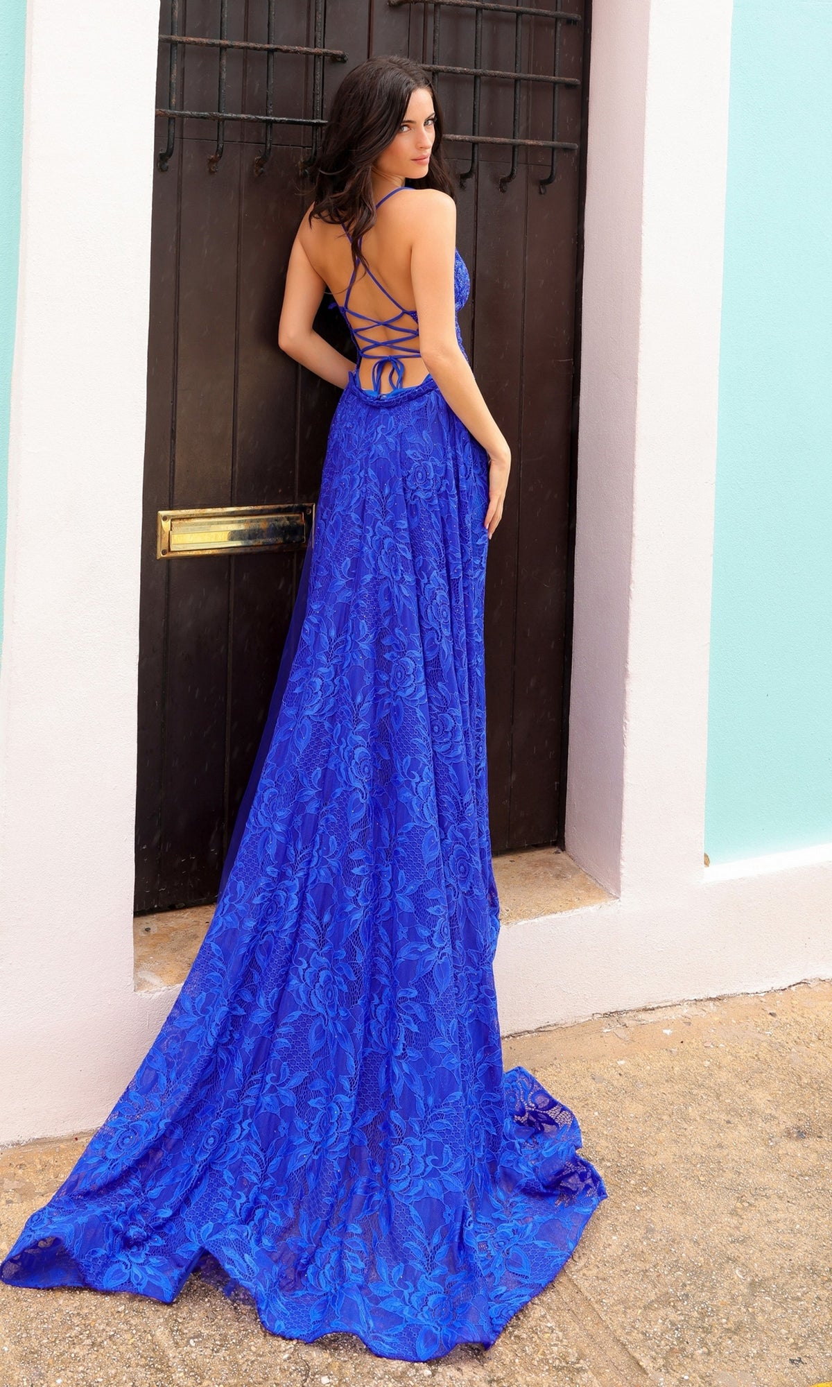 A woman wearing the Open-Back Long Lace Prom Dress with Train G1353 stands in front of a dark wooden door framed by white and pastel blue walls.